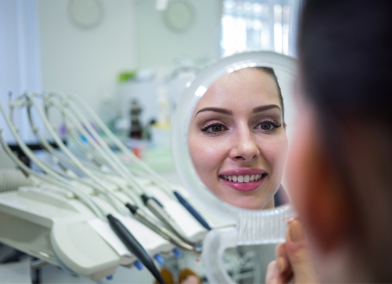 a woman looking herself in a handheld mirror sitting in a dental office